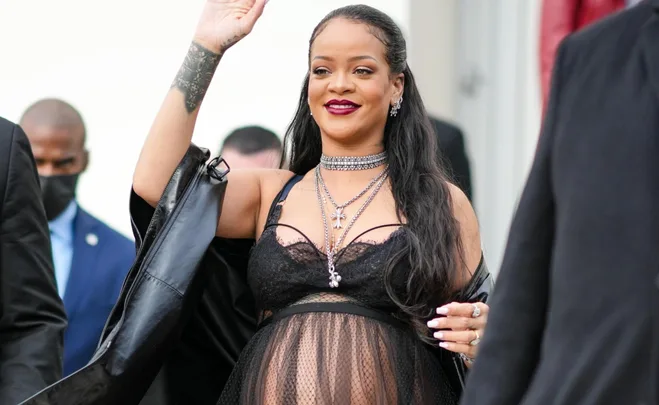 Pregnant woman waves, wearing a sheer black outfit with jewelry, flanked by people in suits.
