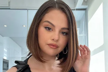 A woman with shoulder-length hair poses in a well-lit room, wearing a black top and hoop earrings.