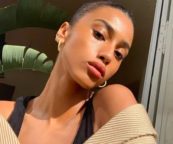 Woman with glowing skin and hoop earrings, posing in bright sunlight near a large green plant indoors.