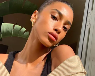 Woman with glowing skin and hoop earrings, posing in bright sunlight near a large green plant indoors.