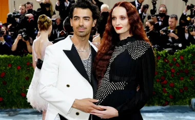 A couple poses on the red carpet, surrounded by photographers and onlookers, with vibrant decorations in the background.