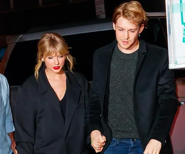 A man and woman walking outside, holding hands, both wearing dark jackets.