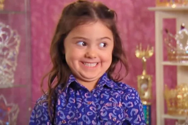 Young girl with long brown hair and a cheeky grin, wearing a blue patterned shirt, stands in a room with trophies.