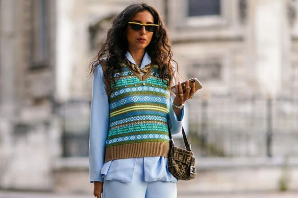 Woman wearing a patterned oversized vest, blue shirt, sunglasses, and holding a smartphone, with a small Fendi bag.