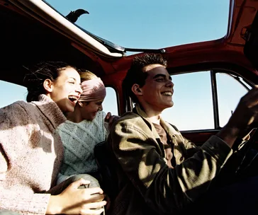 Friends laughing and enjoying a road trip in a car with a clear blue sky outside.