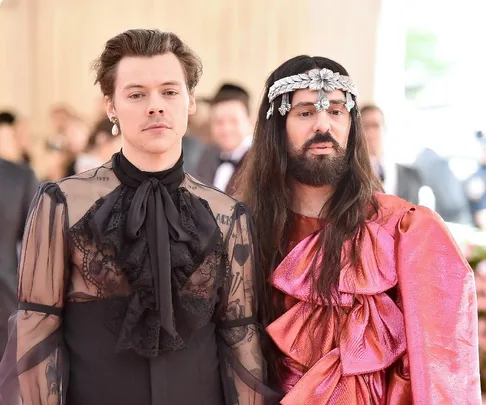 Harry Styles and Alessandro Michele at an event, Styles in a sheer black outfit, Michele in a pink ensemble.