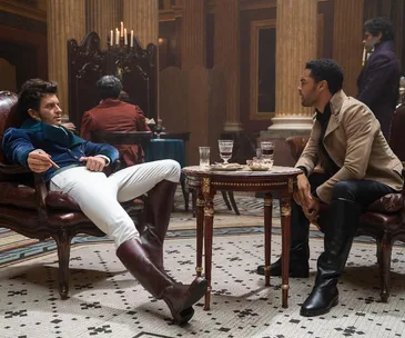 Two men in period costumes sit across a table in an ornate room with drinks and snacks.