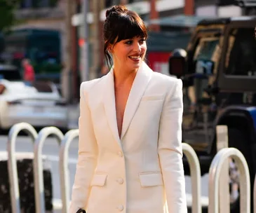 "Person in a stylish white blazer with tied-up hair, smiling outdoors in an urban setting."