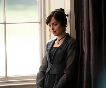 Dakota Johnson as Anne Elliot in period attire, gazing softly near a window, from "Persuasion" film adaptation.