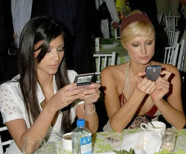 Two women at an event table, focused on their flip phones. Bottled water and cups are on the table.