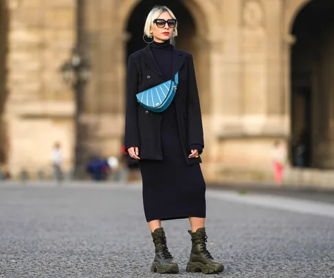 Woman in black coat, long dress, and boots, wears sunglasses and blue crossbody bag, standing on city street.