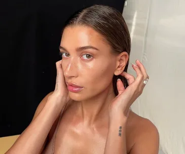 Close-up of a woman with slicked-back hair showing off earrings and a ring, against a neutral background.