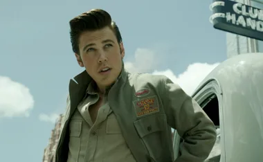 A man with styled hair and a jacket stands next to a vintage car under a "Club Handy" sign, looking into the distance.