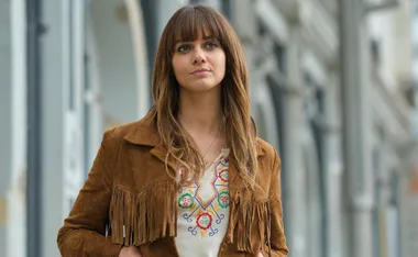 A woman with long brown hair, wearing a brown fringed jacket and a patterned shirt, stands outdoors.