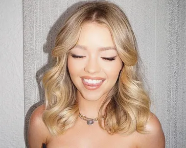 Smiling woman with blonde wavy hair, closed eyes, wearing a necklace and strapless top against a textured wall.