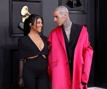 A couple poses at an event, with the woman in a black dress and the man in a pink coat, a Grammy trophy in the background.