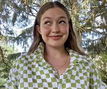 Woman smiling outdoors in a green and white checkered shirt with trees in the background.