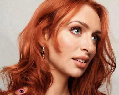 Woman with vibrant red hair and blue eyes gazes to the side, wearing an earring against a neutral background.