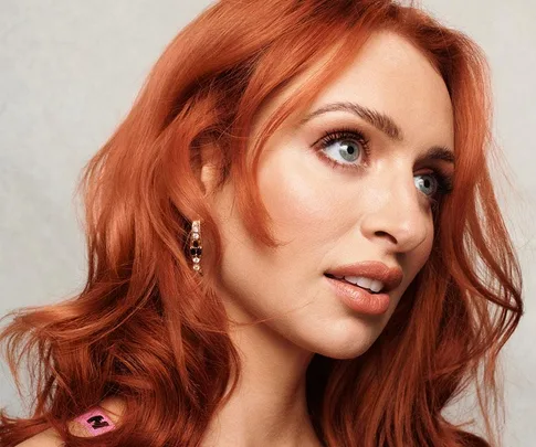 Woman with vibrant red hair and blue eyes gazes to the side, wearing an earring against a neutral background.