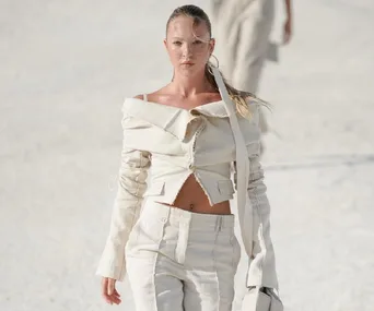 Model wearing an off-shoulder beige ensemble walks confidently on a runway under bright lighting.