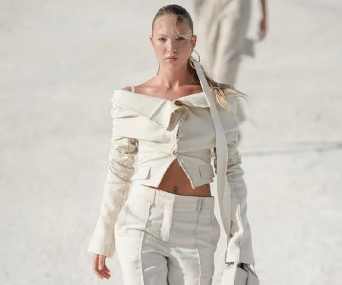 Model wearing an off-shoulder beige ensemble walks confidently on a runway under bright lighting.