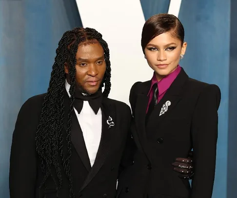 Two people in formal black suits, one with long braided hair, the other with short hair, posing at an event.