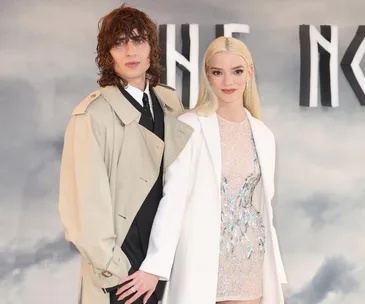 A couple poses on the red carpet, with the woman in a sparkling dress and the man in a trench coat.