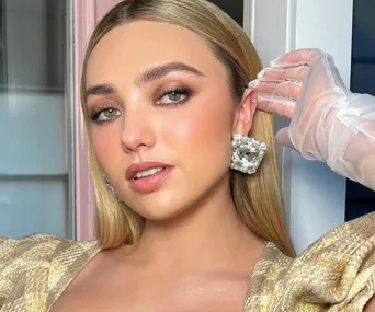 Young woman with long blonde hair, wearing large square earrings and a patterned top, posing indoors with a confident expression.