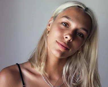 A person with long blonde hair and subtle makeup, wearing a black top and gold earrings, looks softly at the camera.