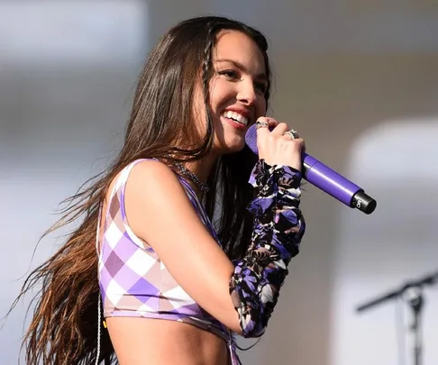 Singer performs on stage with a purple microphone, wearing a checkered top and floral sleeve, smiling passionately.