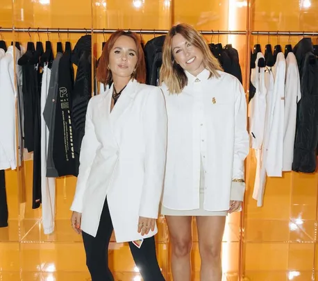 Two women in white outfits posing in a clothing store with racks of jackets in the background.