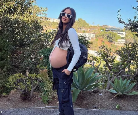 Pregnant woman in a white top and black pants, wearing sunglasses, stands outside on a sunny day.