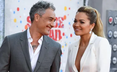 Man in a gray suit and woman in a white dress smiling at each other at a red carpet event.