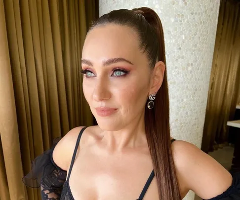 Woman with sleek ponytail and elegant earrings in black dress against a background of brown curtains.