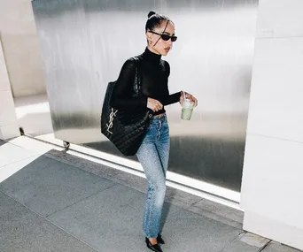 A person in sunglasses walks while holding a green beverage and wearing blue jeans and a Saint Laurent Icare bag.