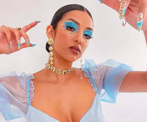 A woman with vibrant blue eye makeup, floral jewelry, nail art, and a sheer blue dress poses with her arms raised.