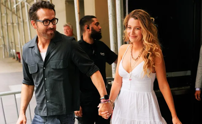 Couple holding hands, woman in a white dress smiles, man in black outfit with glasses; outdoor setting.