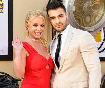 A couple poses together; the woman in a red dress waves, the man in a cream suit stands beside her, smiling at the camera.