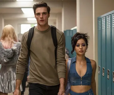 A tall man and a woman with purple eyeshadow walk down a school hallway together, surrounded by lockers.