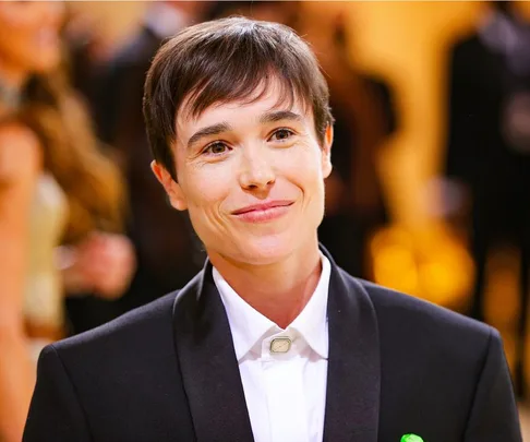 A person with short hair in a black suit smiles at a formal event with a blurred crowd in the background.