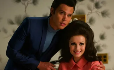 Two actors portray Elvis and Priscilla Presley, with Elvis in blue and Priscilla in pink, in a warmly lit room.