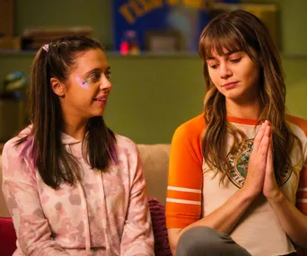 Two women sitting on a couch, one with face glitter smiles at another who has hands together, wearing an orange and white top.