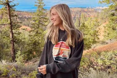 A woman with long blonde hair smiles in a forest, wearing a Zion National Park sweatshirt.