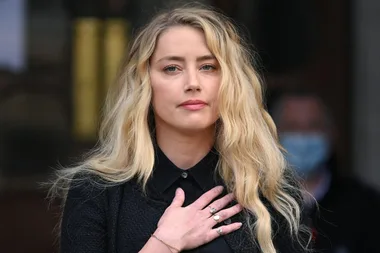 A woman with long blonde hair and a serious expression holds her hand to her chest, standing in front of a blurred background.