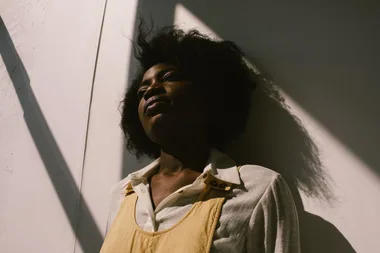 A person with curly hair leans against a sunlit wall, wearing a collared shirt and a mustard-colored dress.