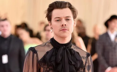 Young man in sheer black outfit with embroidery, wearing a pearl earring, standing in front of a blurred crowd background.