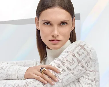 A person with brown hair wearing a patterned white sweater and gold rings, posing with arms crossed.