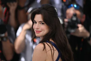 A woman with long brown hair smiles at the camera, surrounded by photographers in a public event.