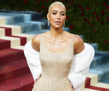 Kim Kardashian in a sparkling nude gown and white fur stole, standing on a red carpet with stairs and greenery in the background.