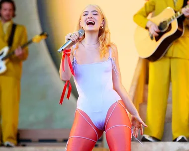 Singer performing on stage with blonde hair, wearing a white outfit, holding a microphone, musicians in background.